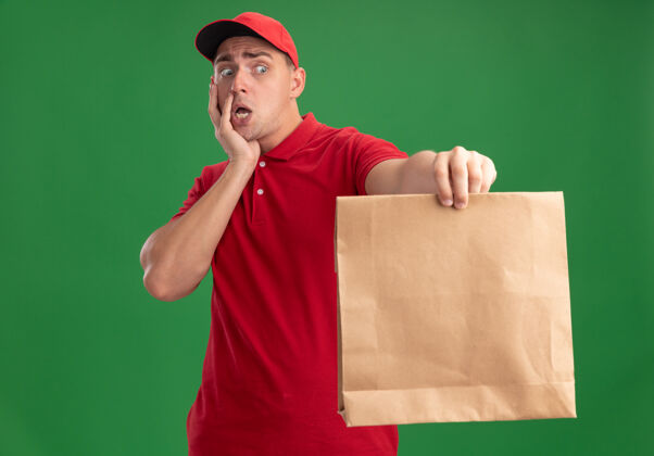 食物年轻的送货员穿着制服 戴着帽子 手里拿着纸制食品包 手放在脸上 被隔离在绿色的墙上送货害怕男人