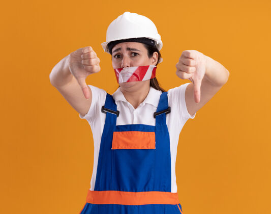建筑工一个穿着制服的年轻建筑妇女 嘴巴被密封 手指朝下 被隔离在橙色的墙上向下女人拇指