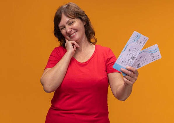 机票身穿红色t恤的中年妇女站在橙色的墙上 手里拿着一张笑脸的机票票位中年女人