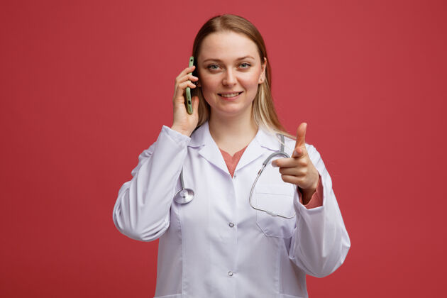 微笑微笑着的年轻金发女医生穿着医用长袍 脖子上戴着听诊器 一边打电话一边看着摄像机女性指指点点金发