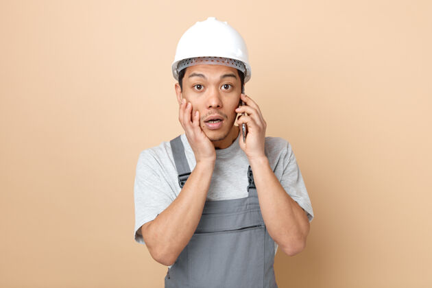 惊喜惊讶的年轻建筑工人戴着安全帽 穿着制服 手贴着脸打电话工人奶油手