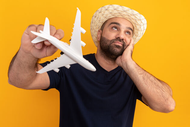 帽子一个留着胡子的男人穿着黑色t恤 戴着夏天的帽子 手里拿着玩具飞机 站在橘色的墙上困惑地抬头看表情飞机人