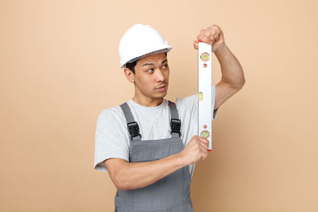 头盔困惑的年轻建筑工人戴着安全帽和制服 拿着水平尺看着施工工人尺子