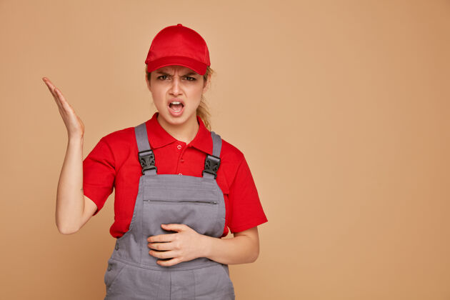 制服愤怒的年轻女建筑工人穿着制服 戴着帽子 手放在肚子上 露出空手女性年轻显示