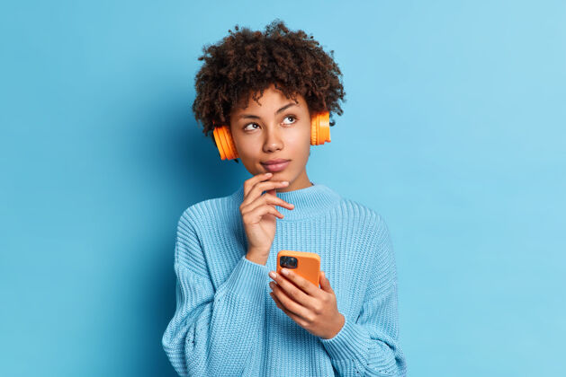 耳机照片中美丽的非洲裔美国女子穿着针织套头衫 用智能手机和脑桥聆听音乐 表情贴心头发毛衣电话