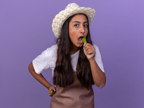 围裙穿着围裙 戴着夏帽 手里拿着青椒的年轻园丁女孩站在紫色的墙上要咬它辣椒站帽子