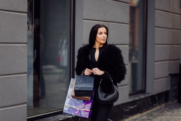 城市一个棕色头发的女人穿着黑色的衣服 手里拿着五颜六色的有图案的购物袋 在一次成功的购物狂欢中走在外面 她享受着一天的温暖女人女性买家