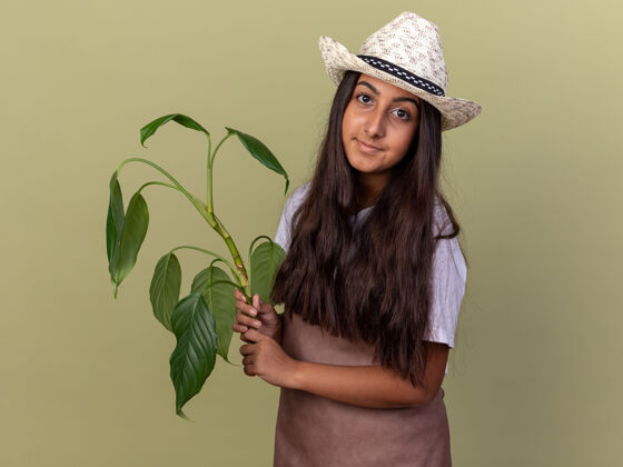 微笑年轻的园丁女孩围着围裙 戴着夏帽 面带微笑地站在绿色的墙上花园围裙年轻