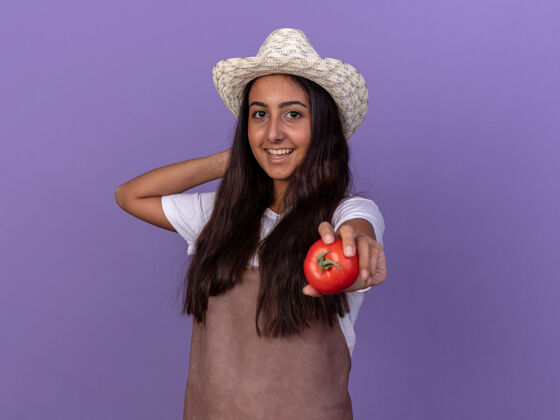 帽子穿着围裙 戴着夏帽 脸上挂着微笑 手里拿着新鲜西红柿的年轻园丁女孩站在紫色的墙上花园微笑站起来