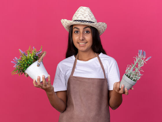 围裙穿着围裙 戴着夏帽 手持盆栽的年轻园丁女孩站在粉红色的墙上 脸上洋溢着幸福的笑容帽子架子罐子