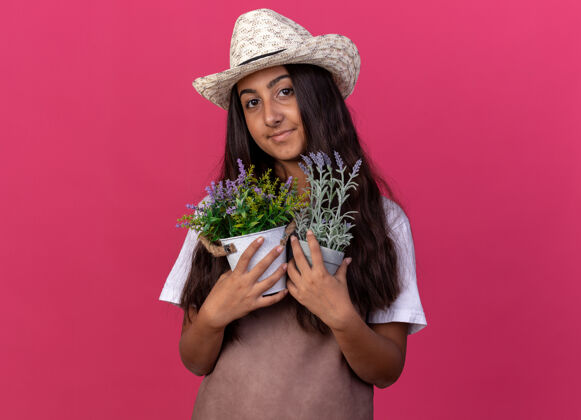 罐子穿着围裙 戴着夏帽 手持盆栽的年轻园丁女孩站在粉红色的墙上 脸上洋溢着幸福的笑容围裙花园年轻的