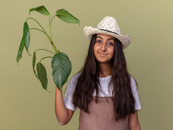 花园年轻的园丁女孩围着围裙 戴着夏帽 抱着植物 自信地微笑着站在绿色的墙上围裙年轻的帽子
