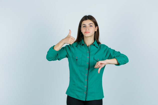 前面年轻的女性展示相反的拇指 弯曲的嘴唇在绿色衬衫 看起来优柔寡断前视图人女性优柔寡断