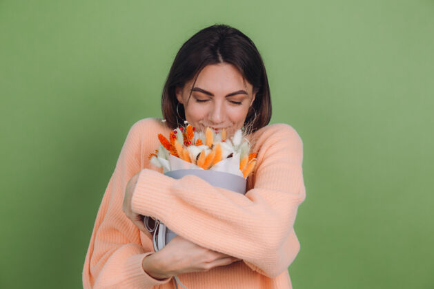 开花年轻女子穿着休闲桃色毛衣隔离在绿橄榄墙上手持橙白色花盒组成的棉花 吉普赛拉小麦和拉古鲁斯作为礼物开心惊喜年轻花束魅力