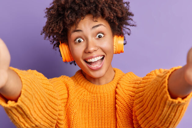 青少年无忧无虑的惊喜快乐的卷曲的非洲裔美国女人睁大眼睛 伸展双手 穿着休闲服自拍 通过立体声无线耳机收听喜爱的音乐民族朋友耳机