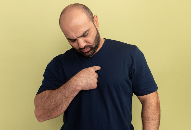 男人一个留着胡子的男人穿着黑色t恤 看起来很困惑 用食指指着站在绿色墙壁上的自己困惑胡须表达