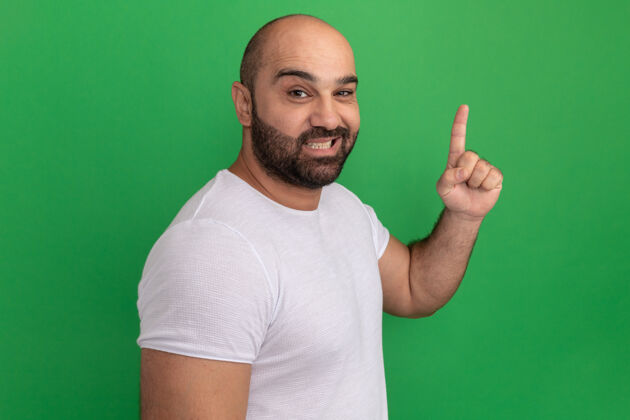 目录快乐的胡子男人穿着白色t恤微笑着伸出食指站在绿色的墙上表演手指胡子