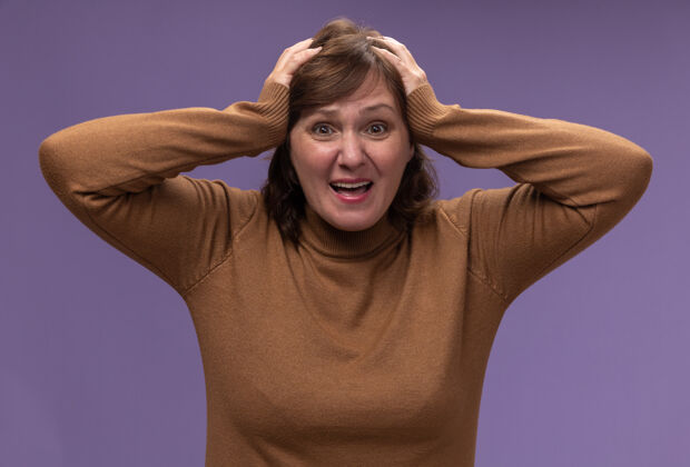 头穿棕色高领套头衫的中年妇女误站在紫色的墙壁上 双手放在头上错误年龄高领毛衣