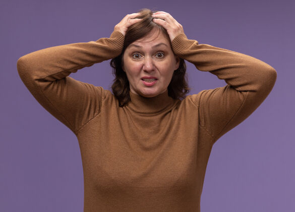 头穿棕色高领套头衫的中年妇女误站在紫色的墙壁上 双手放在头上高领毛衣错误中年
