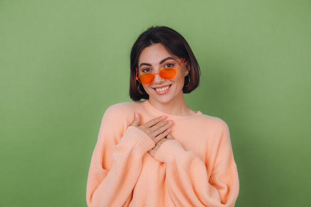 欣赏年轻时尚的女士穿着休闲桃色毛衣 戴着橘色眼镜 隔着绿橄榄色的墙正面双手合十放在胸前 心复制空间可爱人冷静