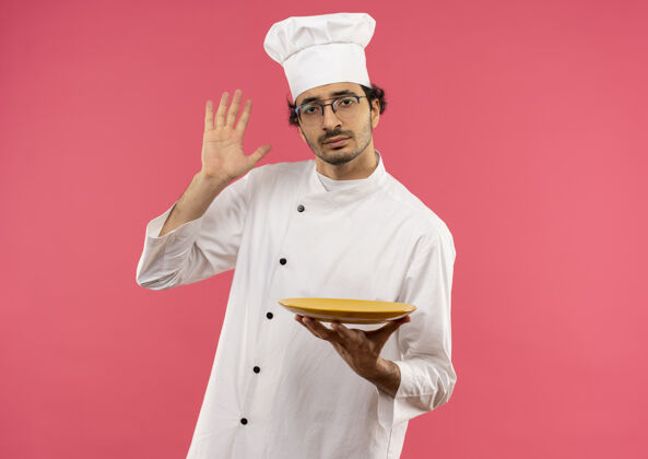 眼镜年轻的男厨师穿着厨师制服 戴着眼镜 拿着盘子举手穿烹饪粉色