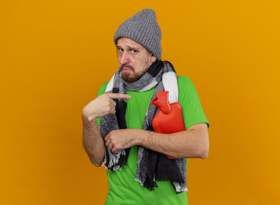 热印象深刻的年轻英俊的斯拉夫病男子戴着冬天的帽子和围巾拿着和指着热水袋孤立在橙色墙上的复制空间斯拉夫年轻穿