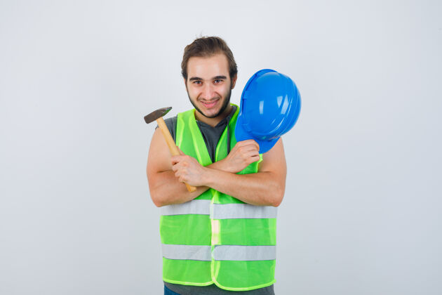 成人年轻的建筑工人手持头盔和锤子 双臂交叉放在胸前 穿着工作服 看上去很高兴正视图男性年轻手臂