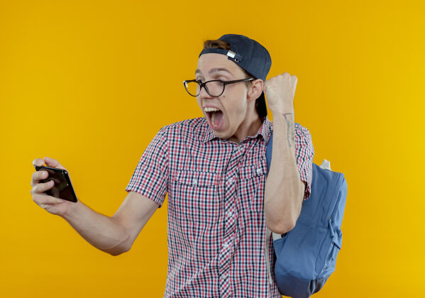 是的快乐的年轻学生男孩背着背包 戴着眼镜 戴着帽子 拿着手机 表示同意戴着拿着手势