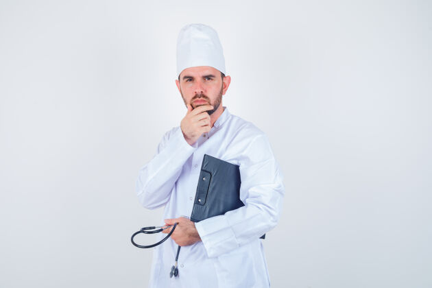 年轻人年轻的男医生拿着剪贴板 听诊器 手放在下巴上 穿着白色制服 看上去很体贴交叉沉思职业