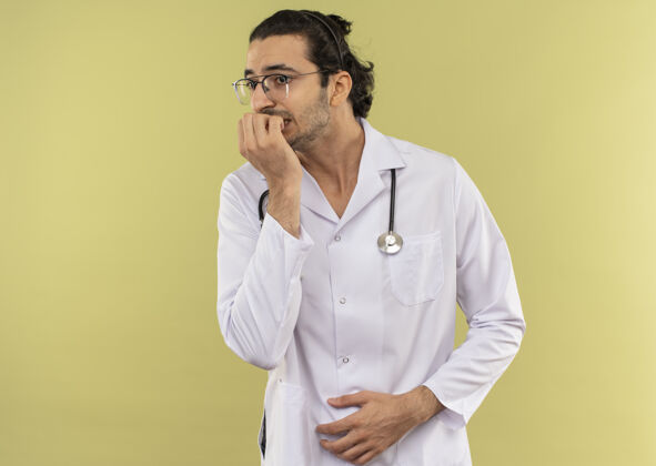 咬关心的年轻男医生戴着眼镜穿着白袍用听诊器咬指甲绿色年轻穿