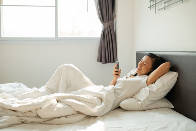 躺下微笑迷人的亚洲女性在白色床上使用平板智能手机女性年轻手机