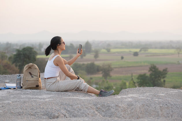 女孩年轻女子拿着手机坐着夕阳下的高山旅游小道复制空间电话举行