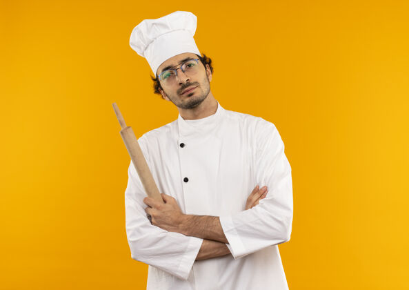 眼镜自信的年轻男厨师穿着厨师制服 戴着眼镜 手里拿着擀面杖 双手交叉滚动交叉黄色