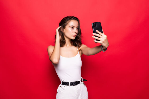 Funky近景写真美女在现代智能手机前自拍隔离在闪亮的红墙上咬年轻微笑