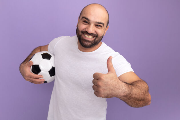 足球快乐的胡子男人穿着白色t恤拿着足球快乐地笑着竖起大拇指站在紫色的墙上表演欢呼男人