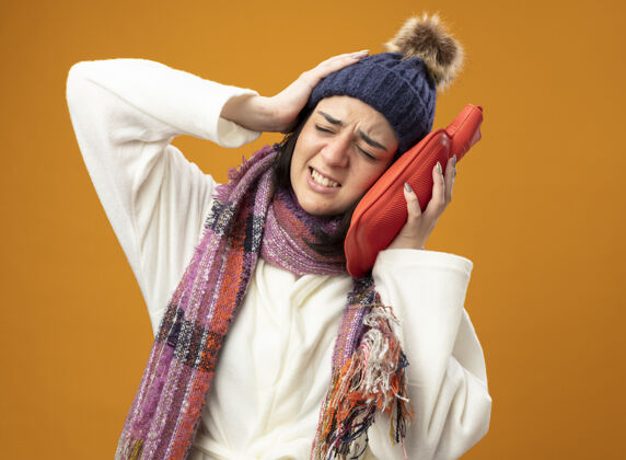 头疼痛的年轻白种人生病的女孩穿着长袍 戴着冬天的帽子和围巾 用热水袋摸着头 手放在头上 闭着眼睛 隔离在橙色的墙上 留着复印空间长袍疾病穿
