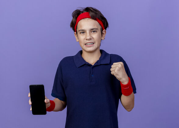 背带微笑的年轻帅气的运动男孩戴着头带和护腕 戴着牙套 显示手机在做“是”的手势 隔离在紫色的墙上 留有复印空间秀腕带男孩