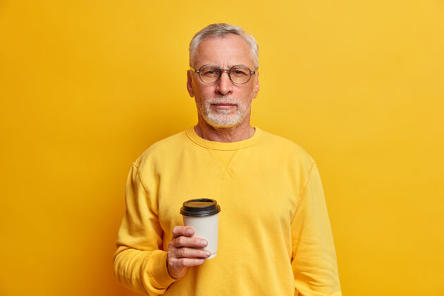 不刮胡子一张孤立的照片 英俊的大胡子男人拿着一次性的外卖咖啡 严肃地看着前面穿着鲜艳的套头衫在黄色的墙上摆姿势休闲手纸