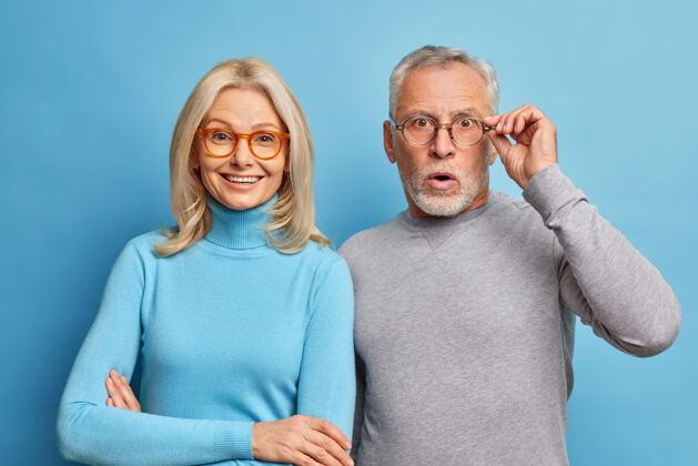 年龄开朗的中年妇女和她的丈夫退休人员对令人震惊的消息作出反应手拿着隔离在蓝色墙壁上的眼镜人肖像老年人