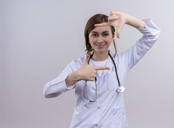 女微笑着的年轻女医生穿着医用长袍和听诊器在隔离的白墙上做着框架手势年轻人女人听诊器