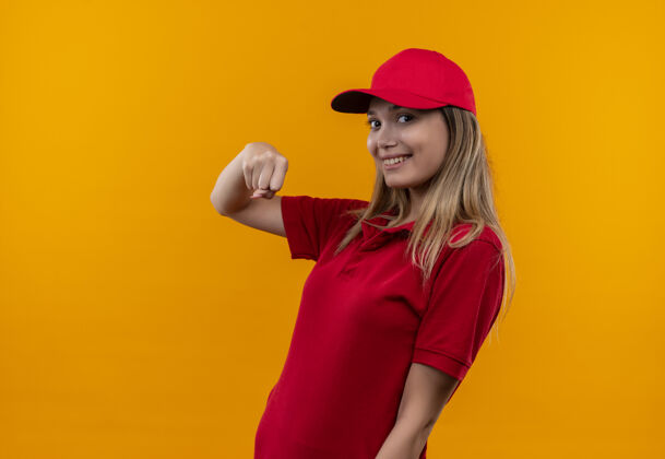 显示微笑的年轻送货女孩身穿红色制服和帽子显示拳头孤立在橙色墙上女孩拳头微笑
