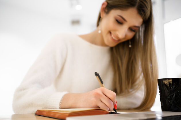 女性微笑美丽的女学生 长发 白色毛衣 在笔记本上写字 同时使用笔记本电脑远程学习远程手机空