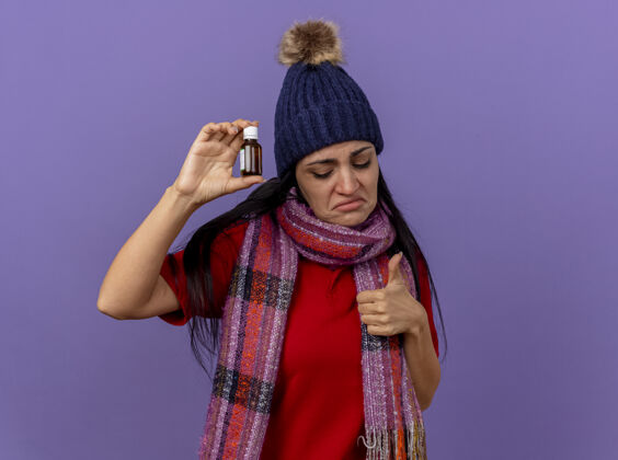 围巾悲伤的年轻白种人生病的女孩戴着冬天的帽子 戴着围巾 手里拿着药水在玻璃里往下看 在紫色的墙上孤立地竖起大拇指 留着复印空间穿疾病显示