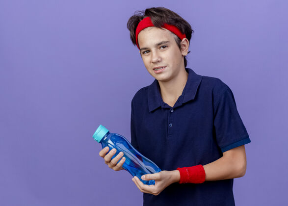 奶瓶高兴的年轻帅气的运动男孩戴着头带和护腕与牙套举行水瓶隔离在紫色墙上复制空间男孩背带头带