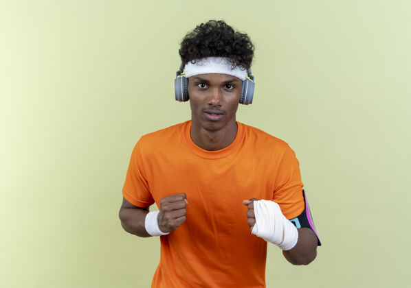 战斗年轻的美国黑人运动男子戴着头带和腕带和电话臂带与耳机站在战斗姿势隔离在绿色的墙壁电话男人立场