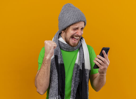 手机快乐的年轻英俊的斯拉夫病夫戴着冬天的帽子和围巾 拿着手机 看着手机做着“是”的手势 隔离在橙色的墙上 留着复印空间疾病斯拉夫橙色