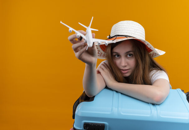 年轻人年轻的旅行女孩戴着帽子伸展模型飞机 把手臂放在手提箱上 放在隔离的橙色墙壁上 留有复制空间手提箱女孩穿