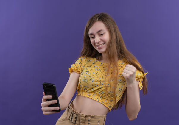 微笑微笑的年轻女孩拿着手机 举起拳头对着孤零零的紫色墙壁年轻电话握着