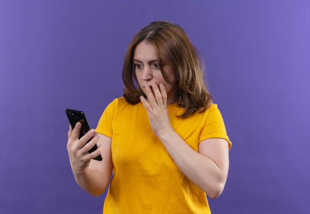 手惊讶的年轻休闲女子拿着手机 用手捂着嘴看着它 在隔离的紫色墙壁上留着复印空间手机年轻休闲