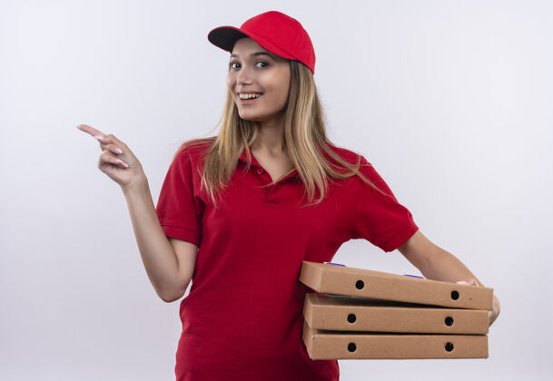 年轻面带微笑的年轻送货女孩身穿红色制服 头戴披萨盒 指着隔离在白色墙上的一侧送货穿女孩